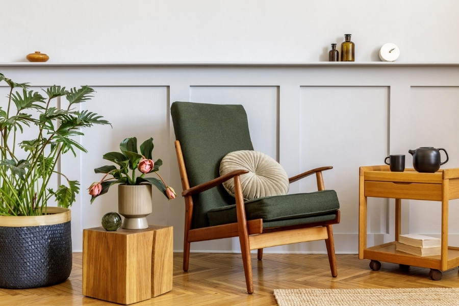 Mid-century fotelja sa zelenim tapacirungom i drvenim rukonaslonima, okružena sobnim biljkama i nameštajem od drveta.
